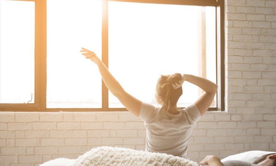 A woman is just waking up, and stretches in front of a set of large custom windows through which the sun is shining.